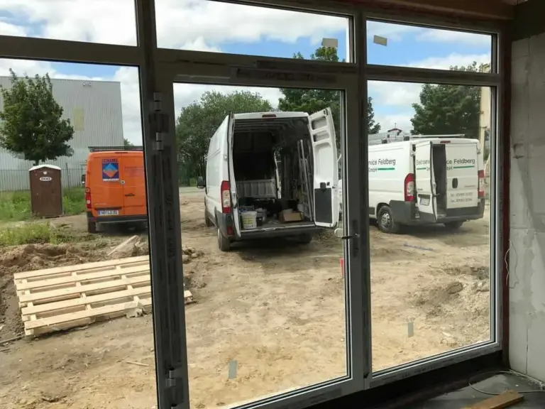 Einbau eines großen Fensters auf einer Baustelle von NORTA. Außenansicht mit geparkten Transportern und Materialpaletten.