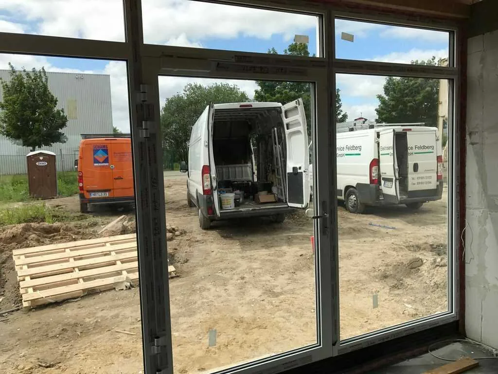 Einbau eines großen Fensters auf einer Baustelle von NORTA. Außenansicht mit geparkten Transportern und Materialpaletten.