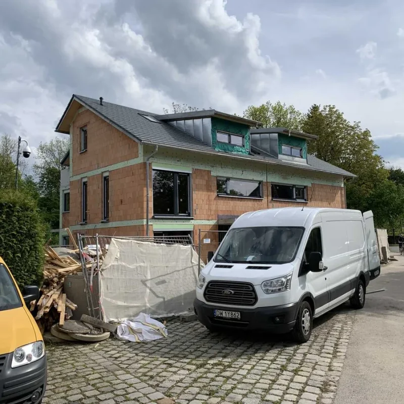 Einfamilienhaus im Bau in München mit weißen Lieferwagen davor, umgeben von Bäumen und Baustellenmaterialien.