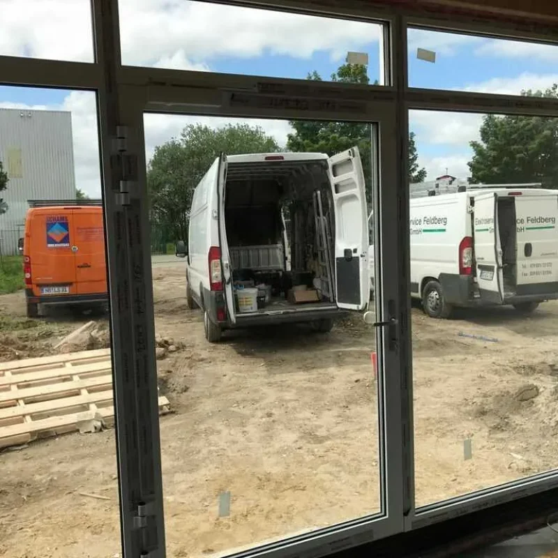 Einbau eines großen Fensters auf einer Baustelle von NORTA. Außenansicht mit geparkten Transportern und Materialpaletten.