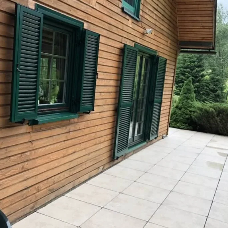 Grüne Fensterläden aus Aluminiumprofilen an einem Holzhaus von NORTA. Witterungsbeständig und dekorativ auf der Terrasse.