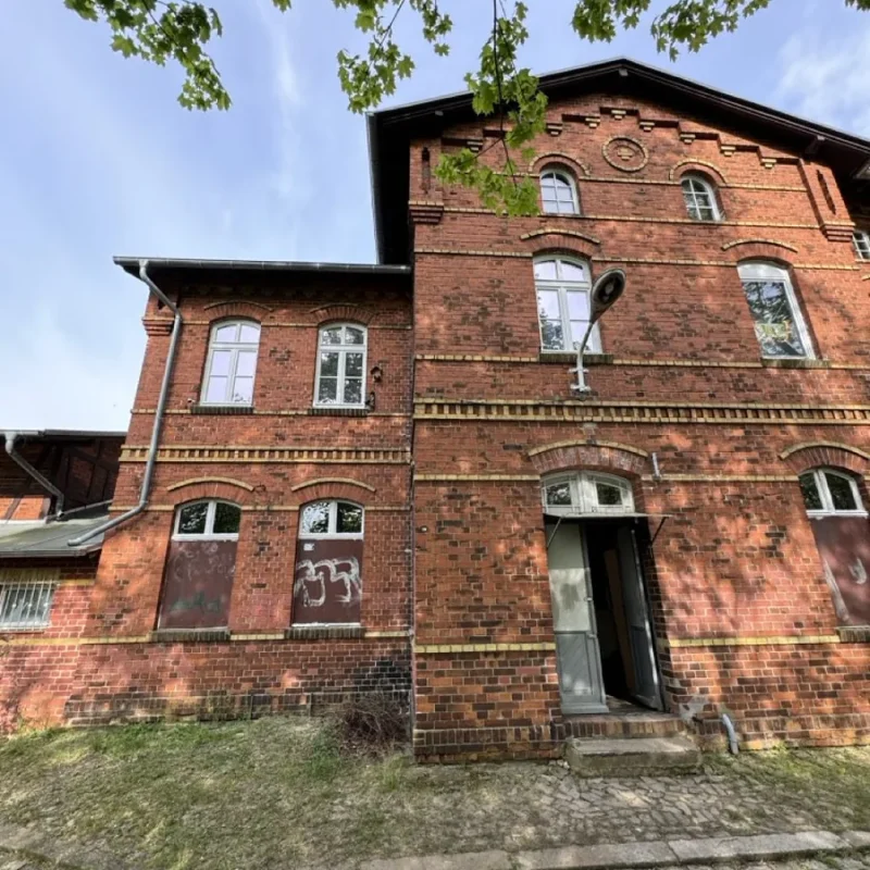 Backsteingebäude mit Holzfenstern und offener Tür, umgeben von Bäumen und Kopfsteinpflasterweg. Hochwertige Lösung von NORTA.