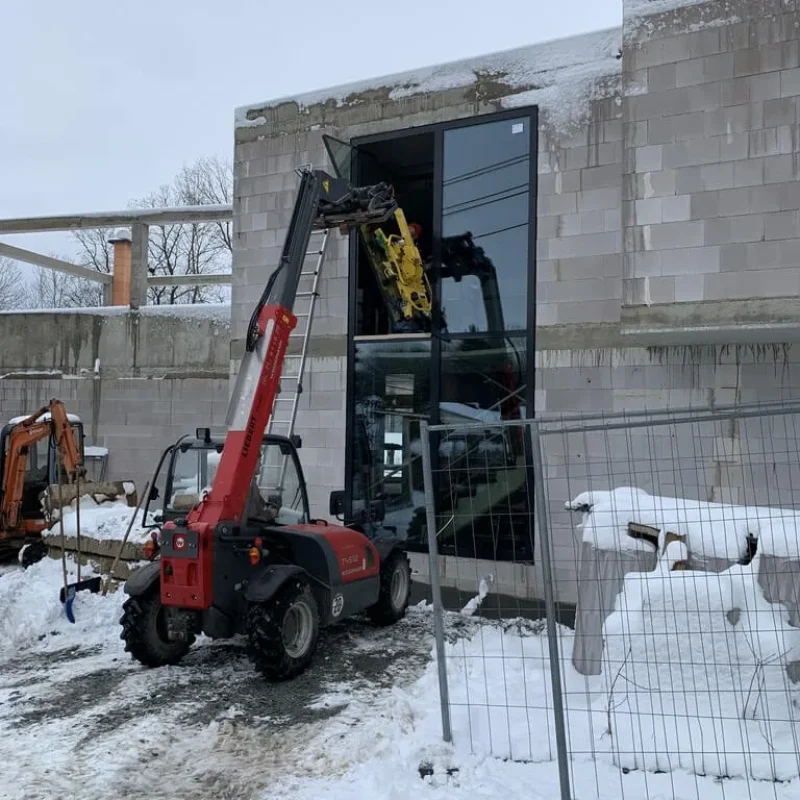 Montage von großem Glasfenster im Winter bei NORTA. Hebemaschine unterstützt den Einbau in einem Rohbaugebäude mit Schnee.